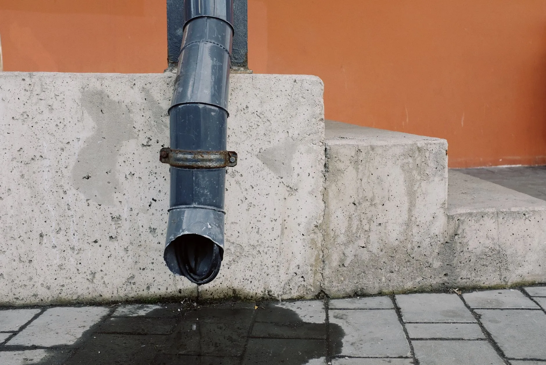 A grey metal downpipe ending above wet paving slabs beside concrete steps