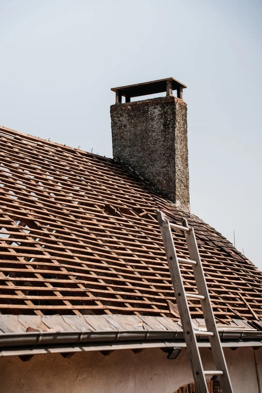 A ladder resting on a tiled roof beside a brick chimney